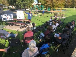 A woman presenting at an outdoor water conservation workshop with attendees seated on chairs in a pa.
