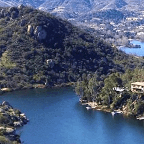 A lush lakeside landscape with hills and residential homes in the background.